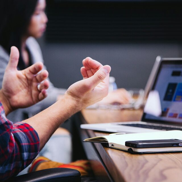 Two colleagues in conversation across a meeting table, one gesturing mid-point